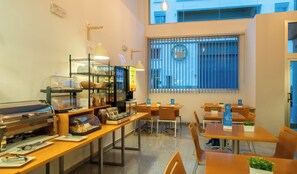 Una cafetería con mostrador de autoservicio, mesas, sillas y un ventanal con persianas.
