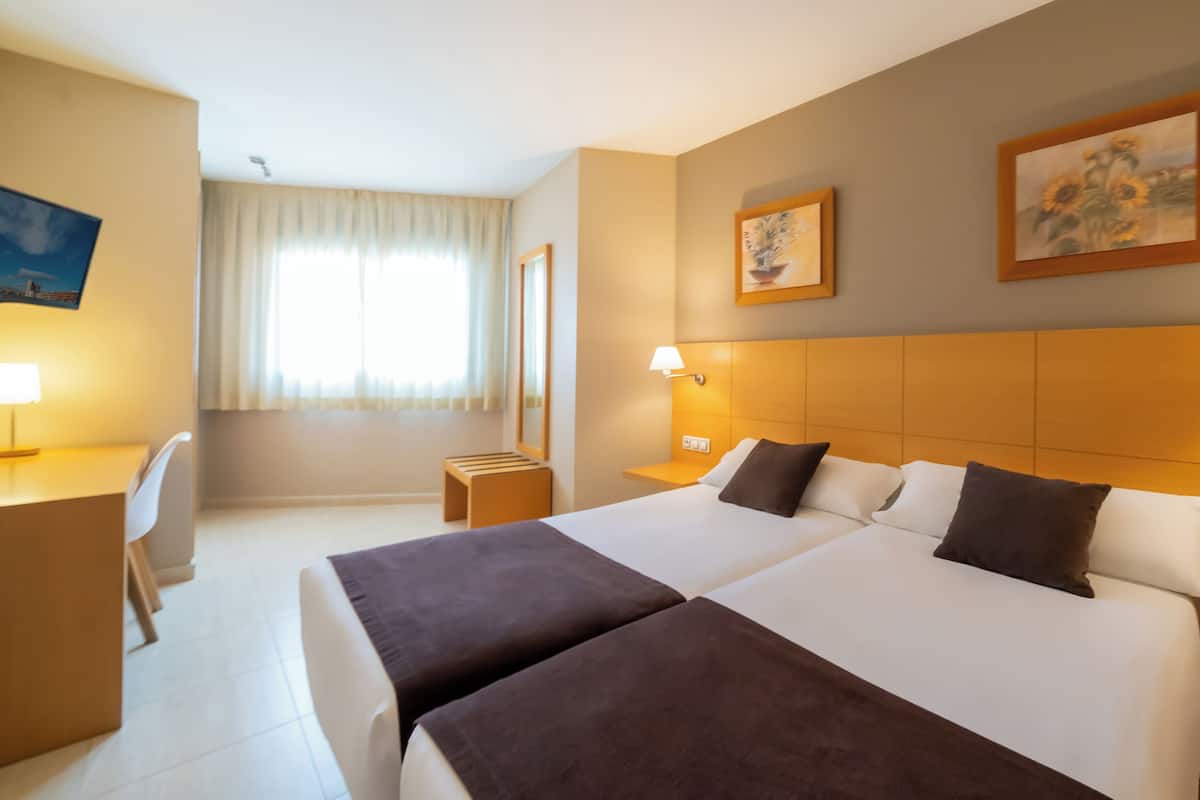 Una habitación de hotel con dos camas, un escritorio, una silla y un ventanal con cortinas.