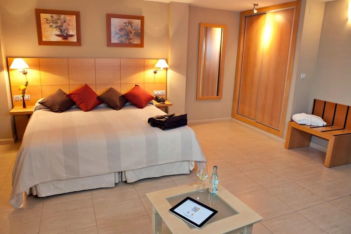 Une chambre d’hôtel avec un grand lit, des lampes de chevet, une table de chevet avec une tablette, un banc en bois et une armoire.