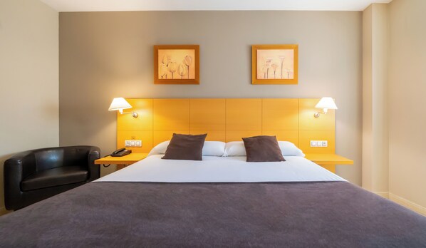 Une chambre d’hôtel avec un grand lit, deux lampes de chevet, une table de chevet avec un téléphone et deux tableaux encadrés au mur.