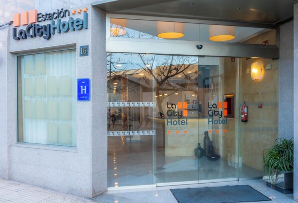 La entrada del Hotel LaCity, con fachada de vidrio, extintor visible y señal de accesibilidad para personas con discapacidad en azul.