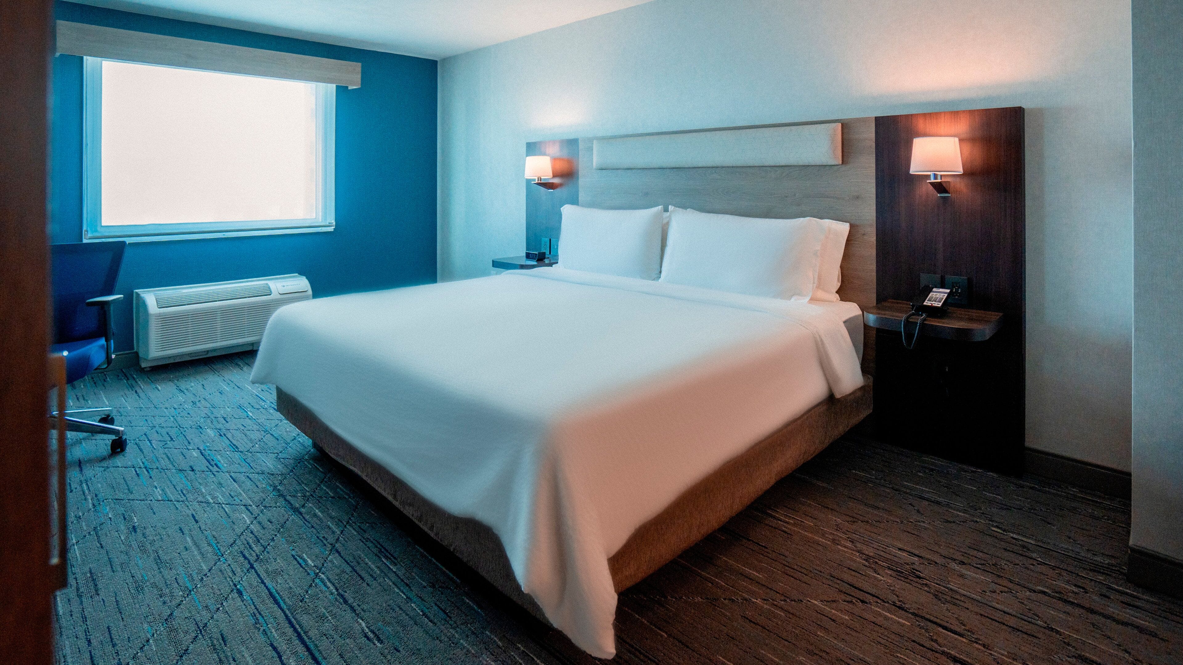 Habitación de hotel con una cama grande, un escritorio con lámpara, una silla, un ventanal y un teléfono en la pared.