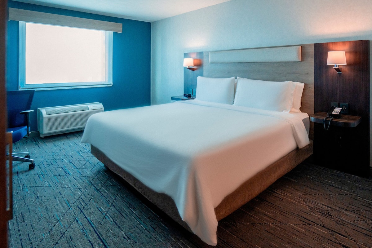 Habitación de hotel con una cama grande, un escritorio con lámpara, una silla, un ventanal y un teléfono en la pared.