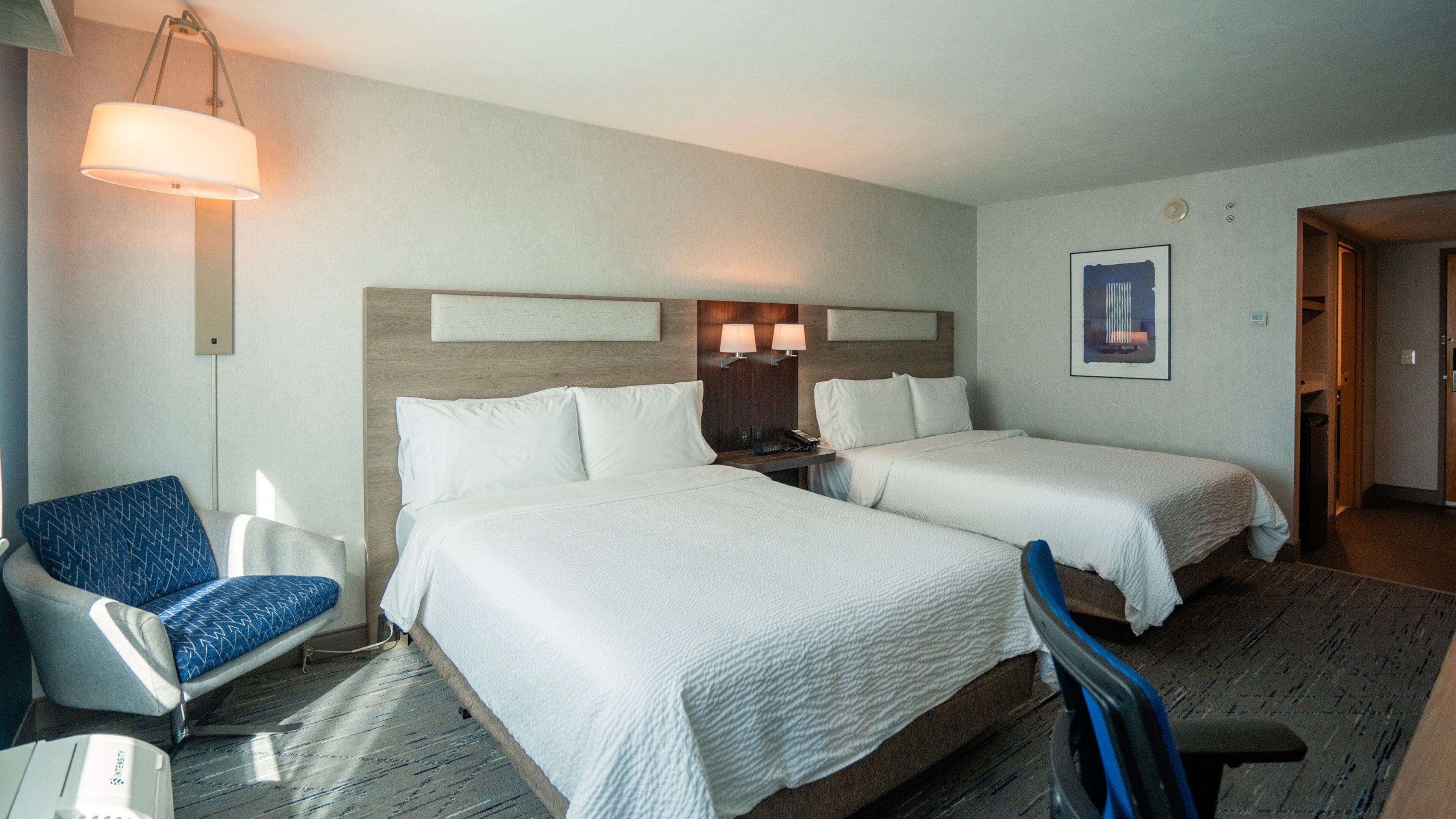 A hotel room with two beds, a desk, a chair, a lamp, and a framed picture on the wall.