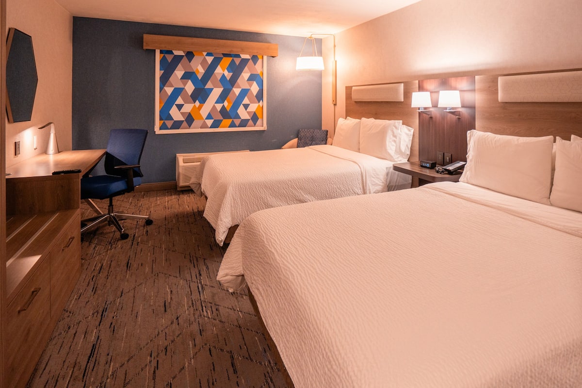 Habitación de hotel con dos camas, un escritorio, una silla, una lámpara de escritorio, un televisor montado en la pared y un tapiz con motivos estampados.