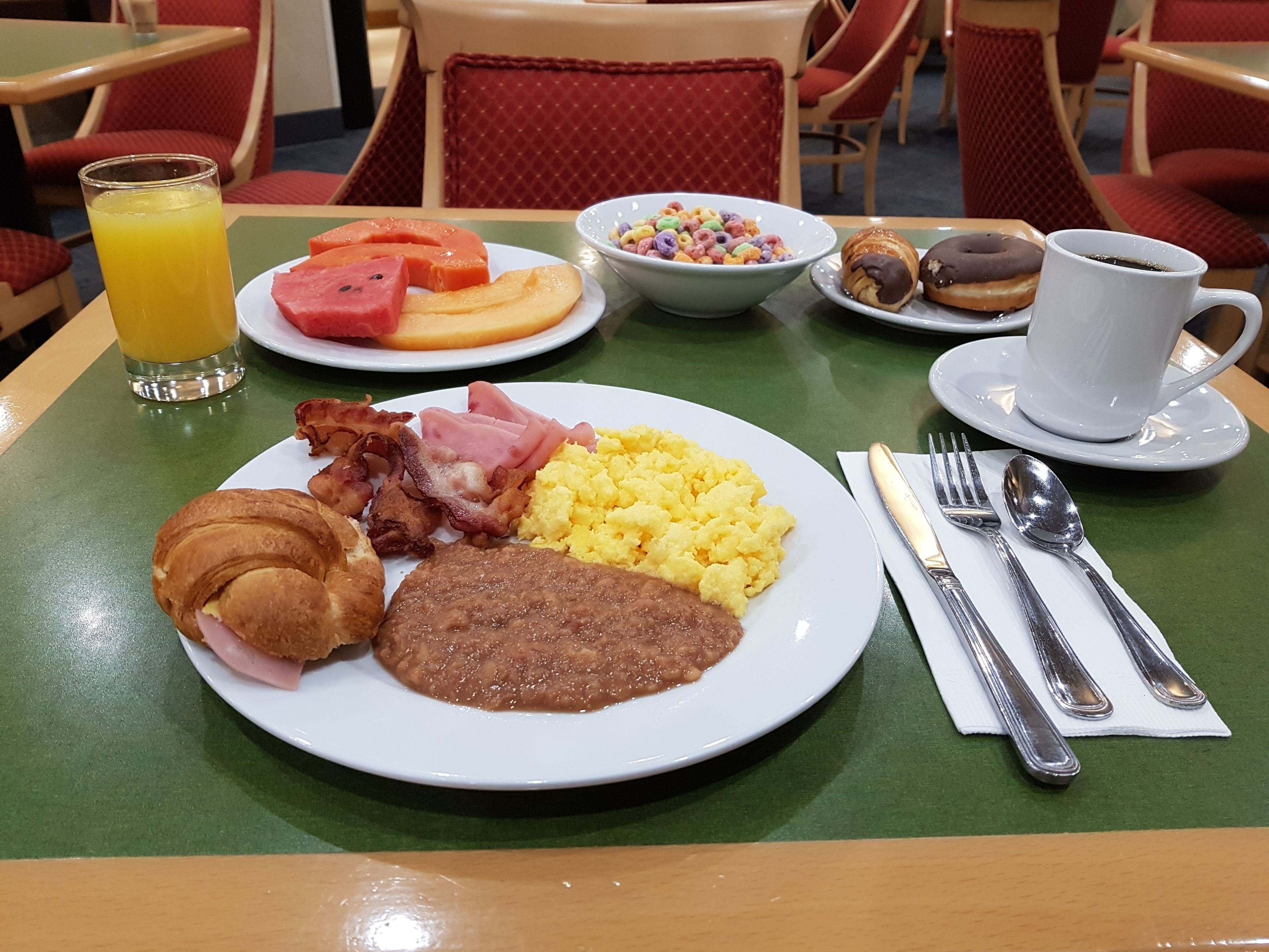 Un desayuno con un plato de huevos, tocino, jamón y un croissant, un tazón de cereal, un vaso de jugo de naranja y una taza de café sobre una mesa.