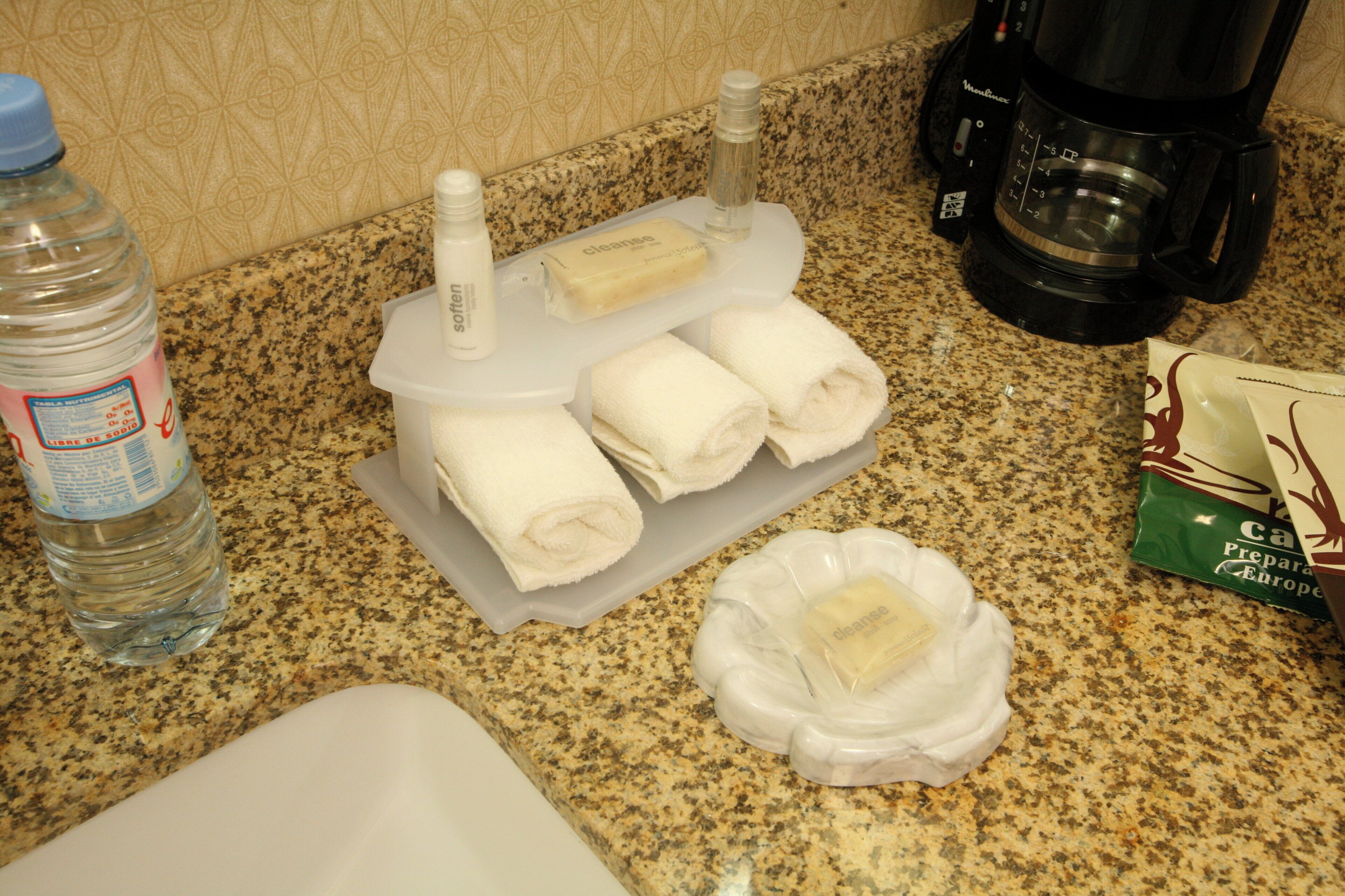 Un comptoir de cuisine avec une bouteille d’eau, une machine à café, un flacon de liquide vaisselle, un savon et un paquet de crème à café.