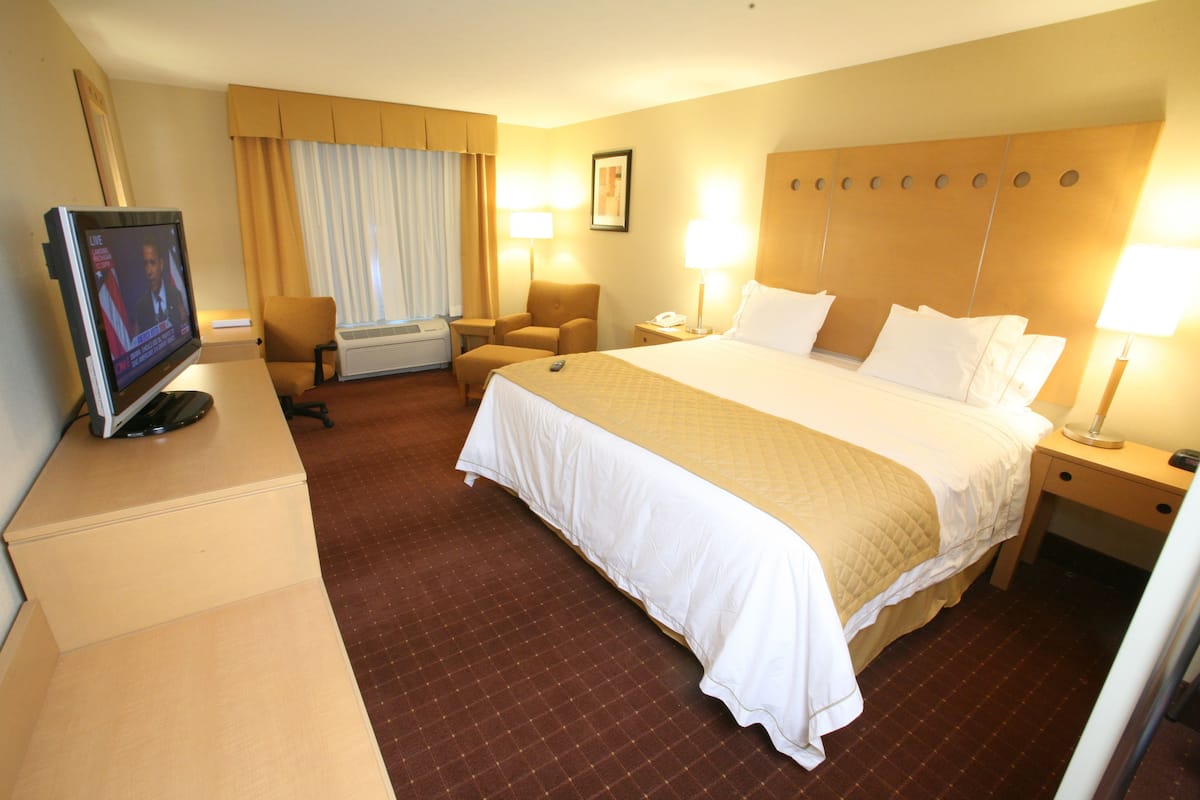 A hotel room with a large bed, a desk with a television, a chair, a nightstand with a lamp, and a window with curtains.