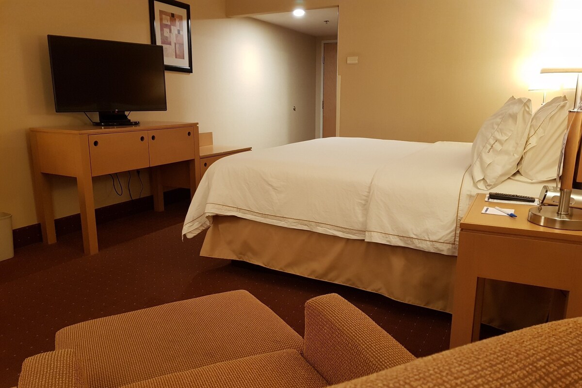 A hotel room with a bed, a television on a wooden cabinet, a desk with a lamp, and a chair.