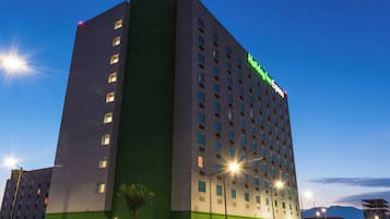 Un hotel de varios pisos con fachada verde y blanca, múltiples ventanales y un letrero visible de Holiday Inn Express.