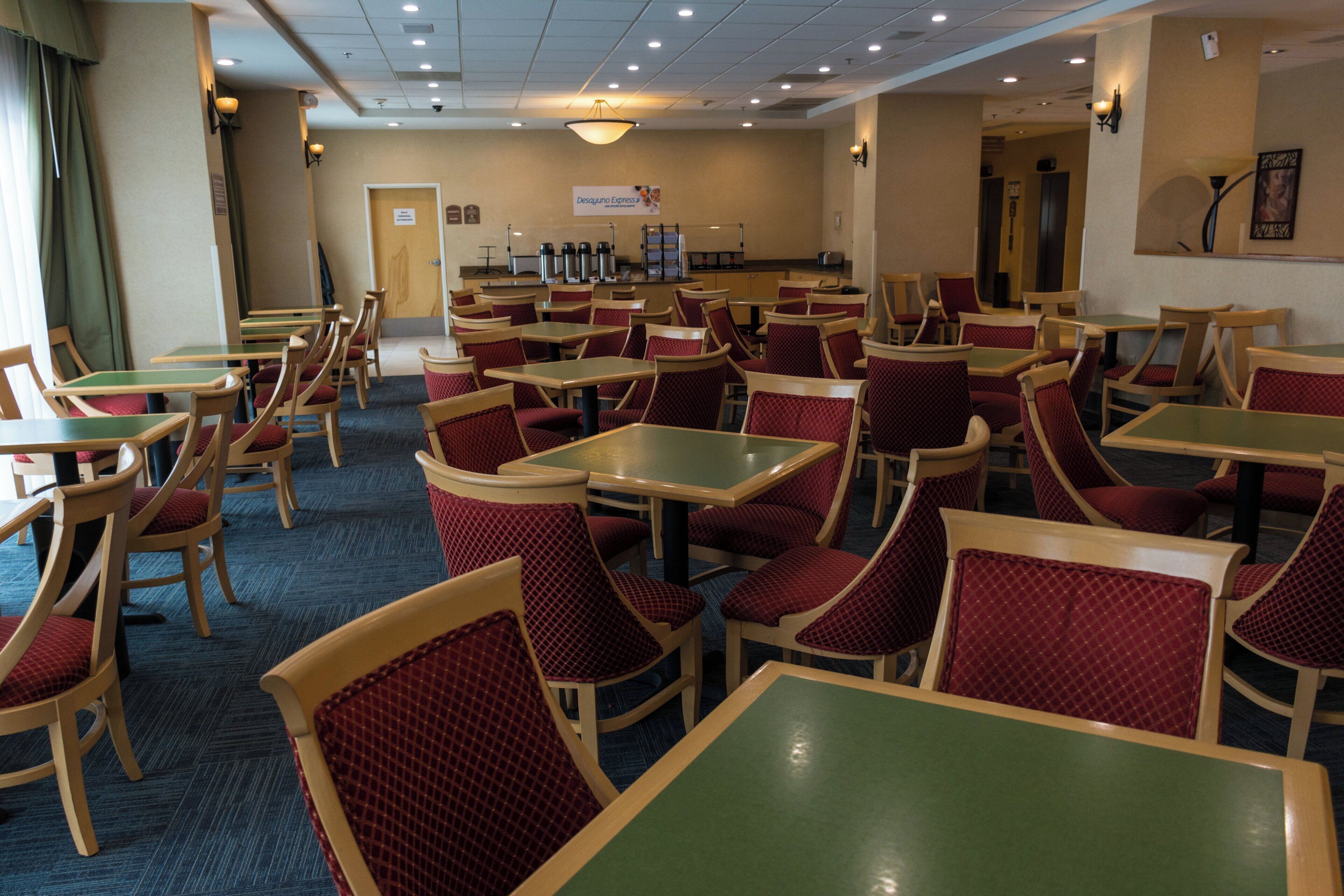 A hotel dining area with multiple tables and chairs, a kitchen in the background, and a sign for Desayuno Express.