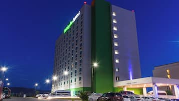 Un hotel de varios pisos con una fachada llamativa en blanco y verde, un estacionamiento con varios coches y un cielo despejado.