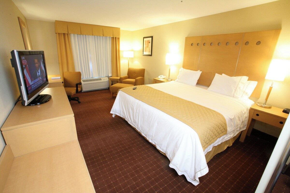 A hotel room with a large bed, bedside lamps, a desk with a television, a chair, and an air conditioning unit.