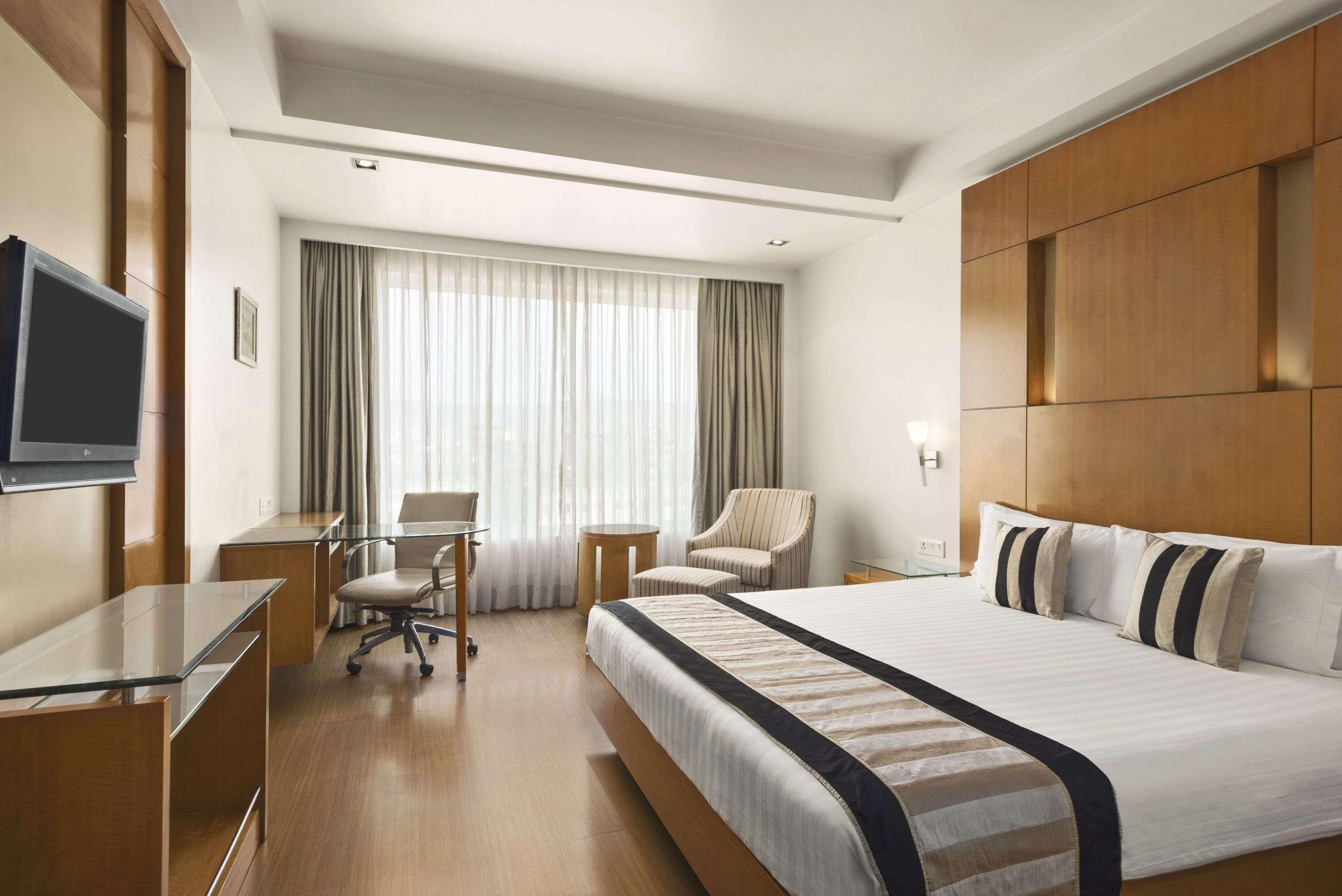 Quarto de hotel com cama, escrivaninha, cadeira, televisão e janela com cortinas.