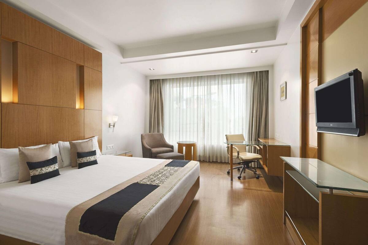 Quarto de hotel com uma cama grande, uma escrivaninha, uma cadeira, uma televisão e uma janela com cortinas.