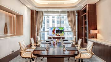 Un salón de conferencias con una gran mesa, sillas, una pantalla de televisión y vistas a edificios a través de amplias ventanas.