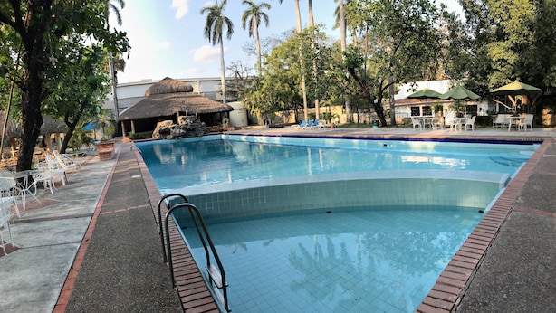 A swimming pool with a tiled edge, surrounded by lounge chairs and umbrellas, with trees and a building in the background. - Hotel Valles (Ciudad Valles)