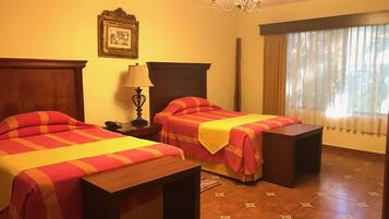 Habitación de hotel con dos camas, una mesita de noche, una lámpara, un cuadro en la pared y una ventana con cortinas.