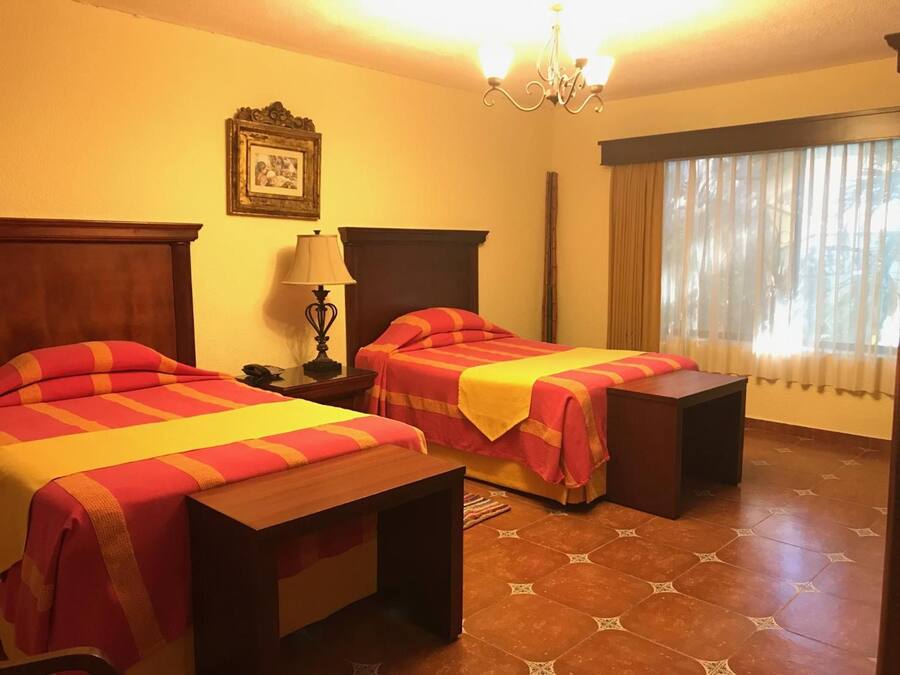 Habitación de hotel con dos camas, una mesita de noche, una lámpara, un cuadro en la pared y una ventana con cortinas.