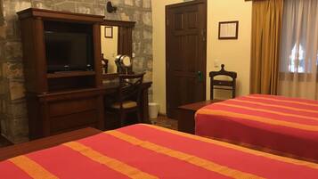 A hotel room with a stone wall, a television, a desk with a mirror, two beds with red and orange striped bedding, and a door marked "SALIDA DE EMERGENCIA".