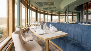 Un restaurante con una larga mesa dispuesta para comer, asientos tapizados en azul y amplias ventanas que ofrecen vistas.