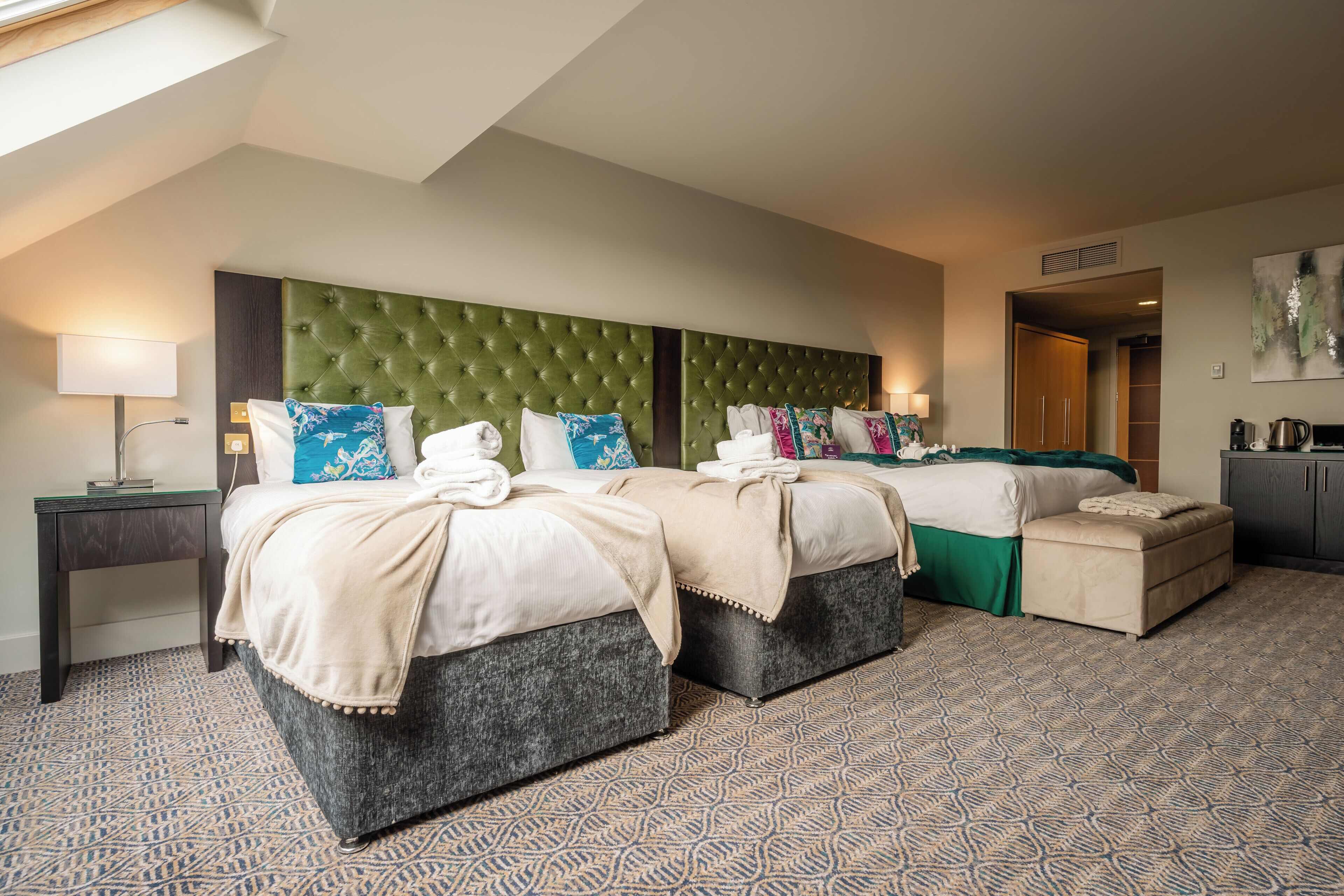 A hotel room with two beds, a green tufted headboard, patterned carpet, and a kitchenette.