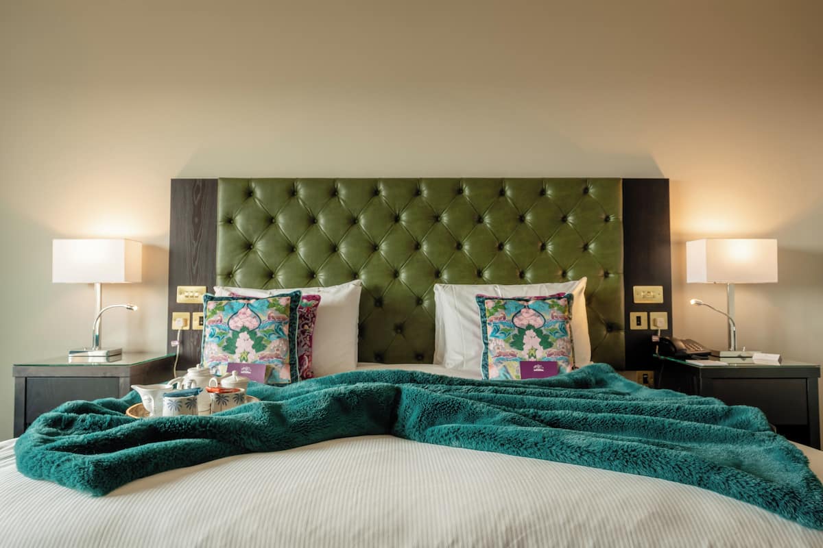 Una cama bien hecha con un cabecero verde, dos lámparas de noche y almohadas estampadas con motivos florales.