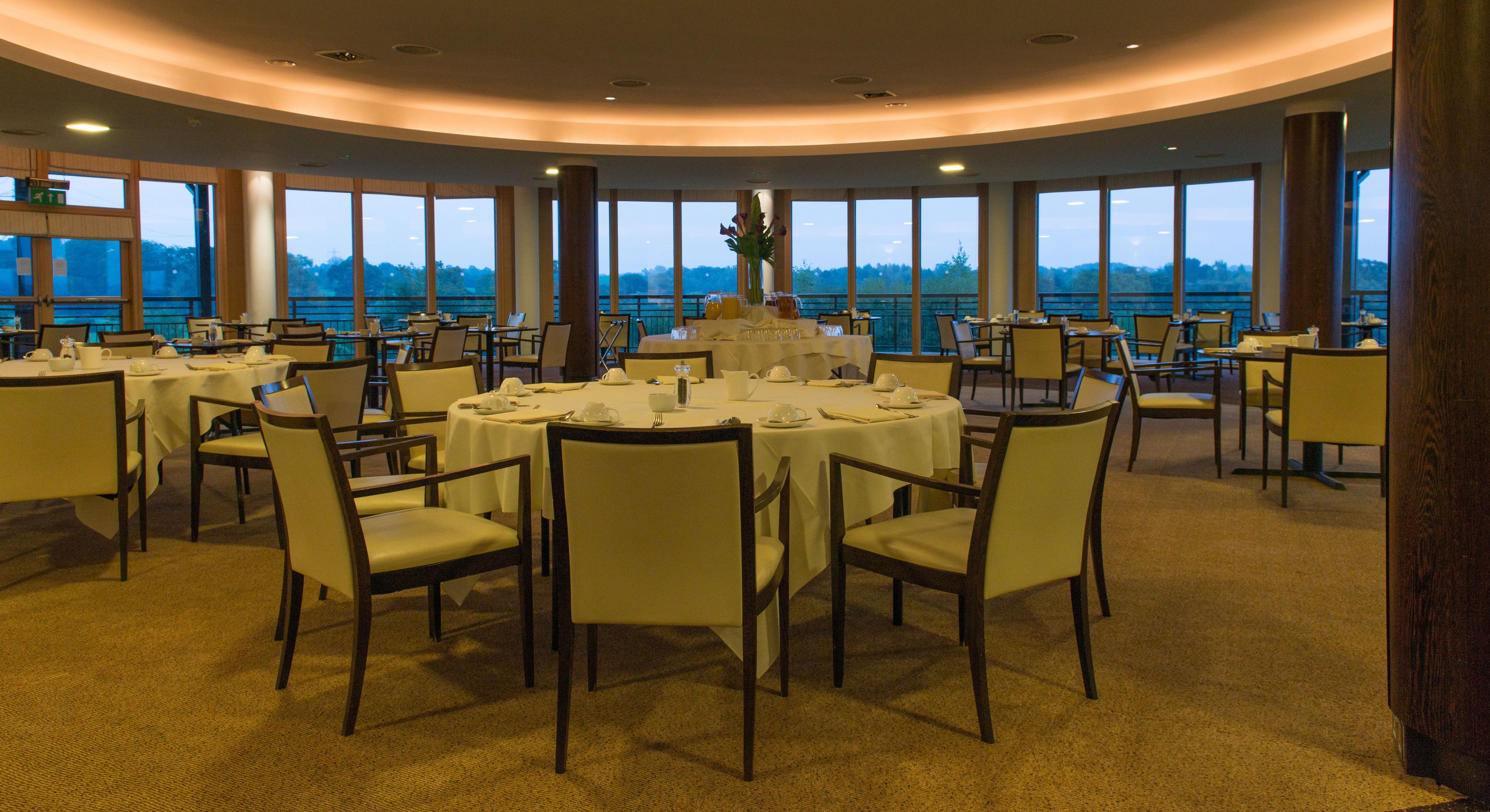 A large, circular dining area with round tables and chairs, large windows offering a view of the outdoors, and a high ceiling with recessed lighting.