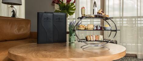 Une table ronde en bois, un canapé en cuir noir, une lampe, un vase avec des fleurs et un plateau contenant des sandwichs et des pâtisseries.