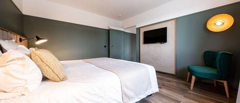 A modern hotel room with a large bed, a desk, a chair, a flat-screen TV, and a wall-mounted lamp.