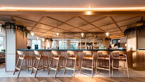 Un espace bar moderne avec un comptoir en bois, de nombreux tabourets de bar, des luminaires suspendus et des étagères où sont exposés des verres.