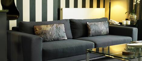 A modern living room with a grey sofa, two patterned cushions, a glass coffee table, and a lamp.