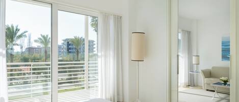 A hotel room with a large bed, a floor lamp, a view of palm trees and buildings through the window, and a door leading to another room.