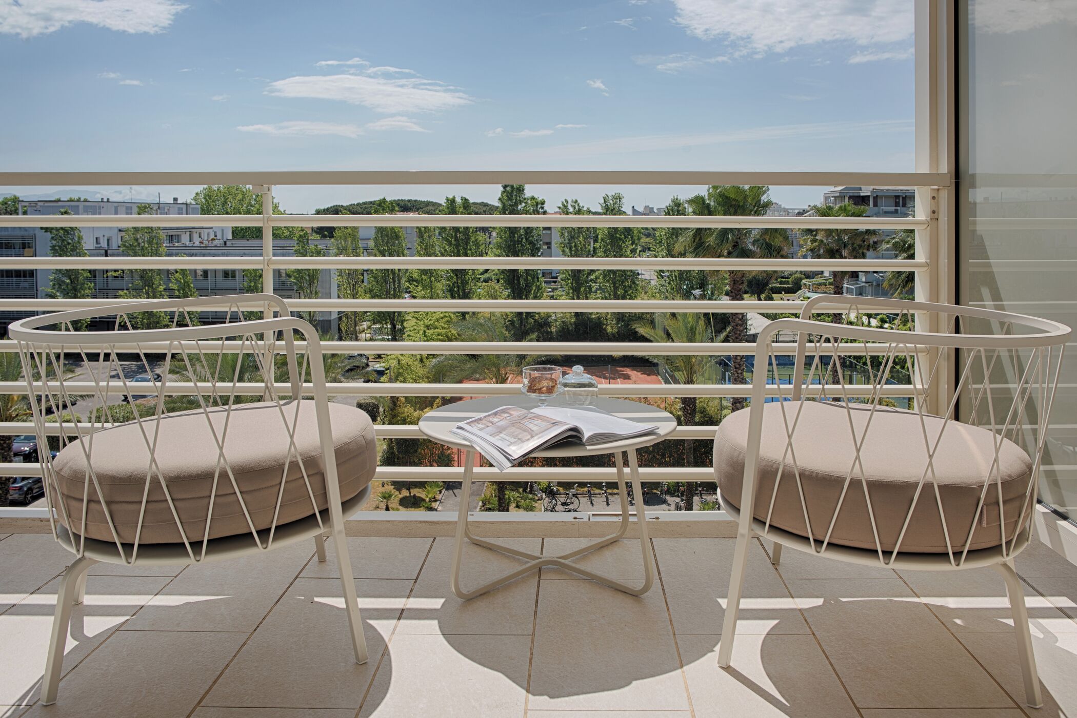 Een balkon met twee comfortabele stoelen, een tafeltje met een tijdschrift, en uitzicht op gebouwen en groen.