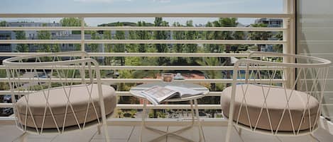 Un balcón con dos sillas acolchadas, una mesita con una revista y vistas a edificios y vegetación.
