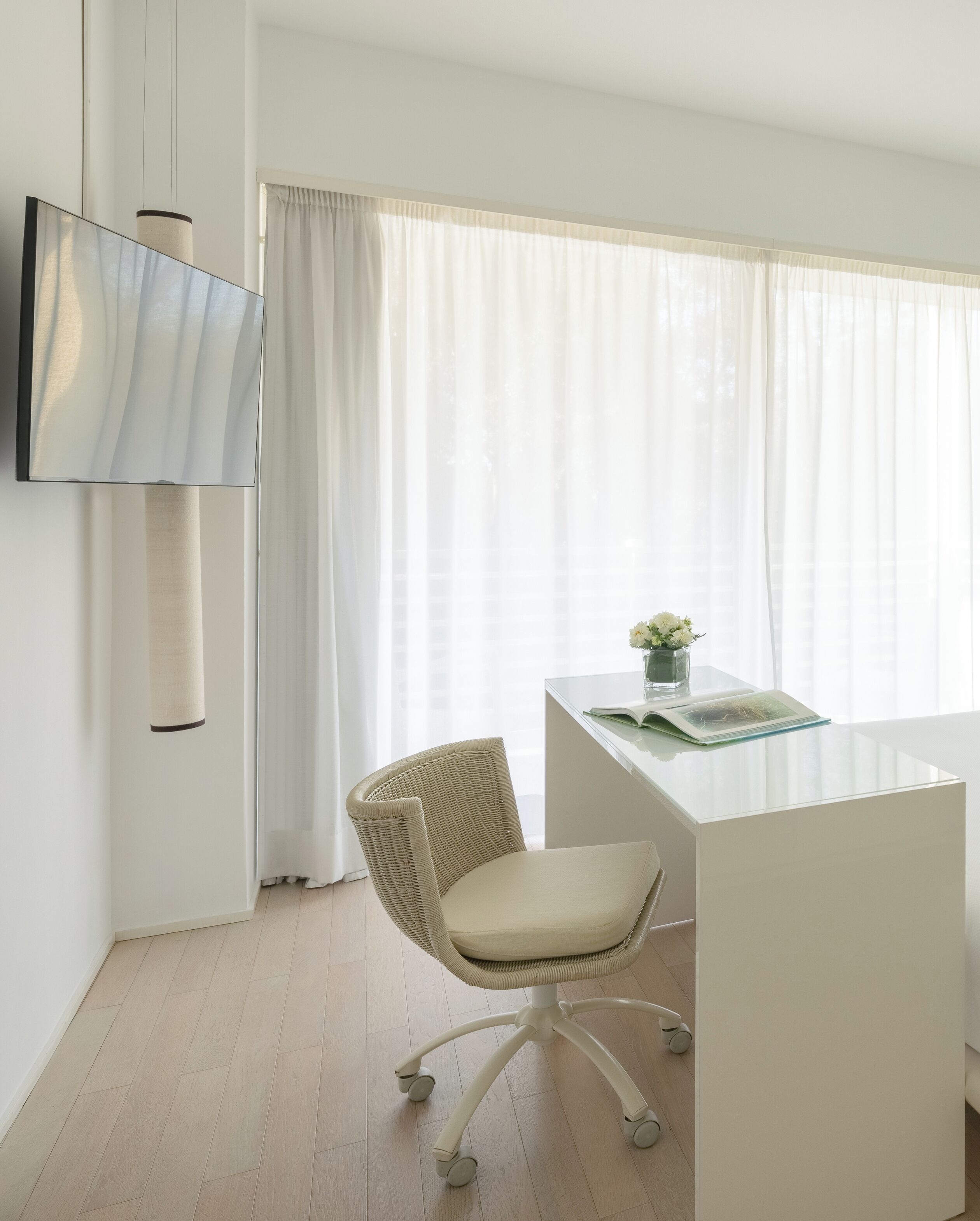 A modern office space with a white desk, a wicker chair, and a large window with sheer curtains.
