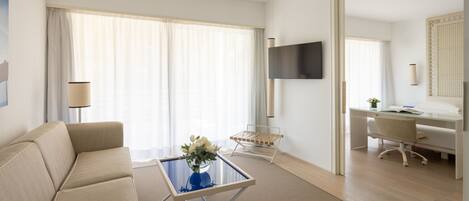 Una habitación de hotel moderna con sofá, una mesa de centro de vidrio con flores, un escritorio con silla y un televisor montado en la pared.