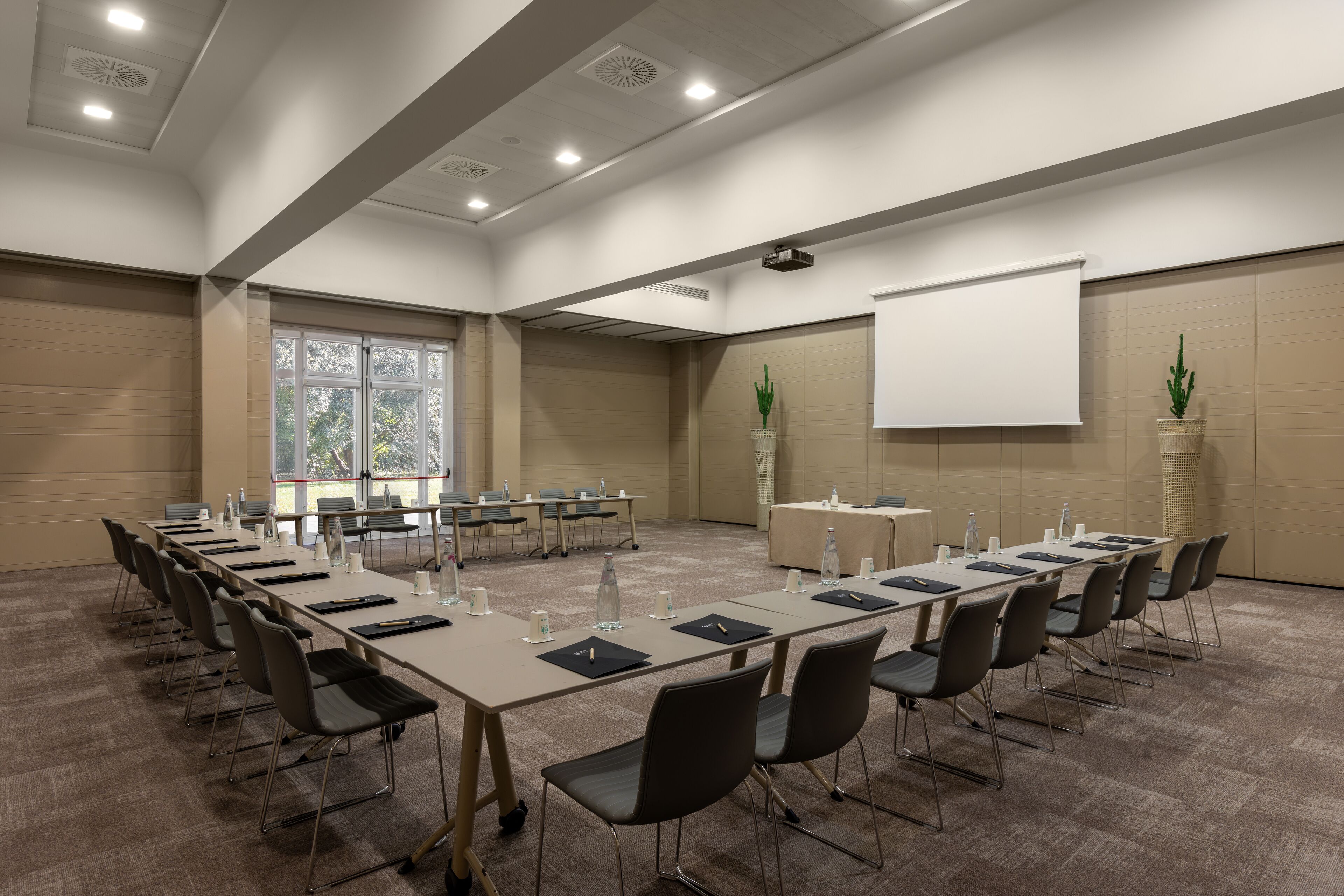 Una sala conferenze con un lungo tavolo, sedie, uno schermo per proiettore e piante in vaso.