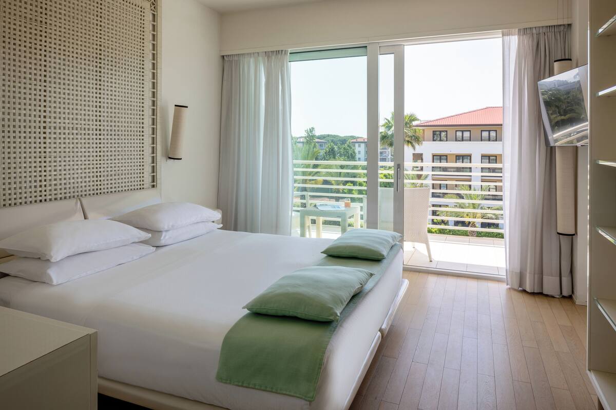 A hotel room with a large bed, a balcony with a view of buildings and greenery, a TV mounted on the wall, and a window with curtains.