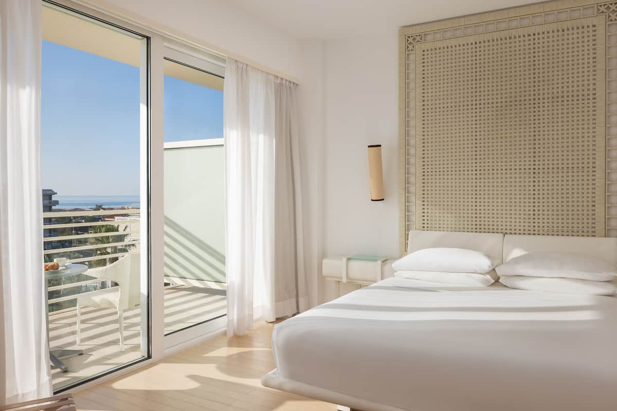 A hotel room with a large bed, a view of the city through a balcony, and a large window with white curtains.
