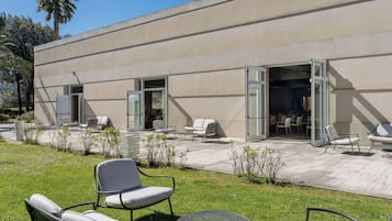 Un espacio al aire libre con sillas y una mesa frente a un edificio con puertas abiertas.