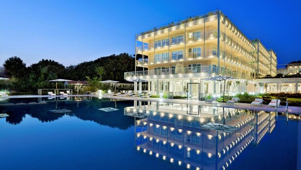 Un hôtel à plusieurs étages avec une piscine en premier plan, doté d’une barrière en verre et de chaises longues.