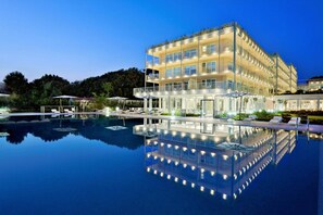 Un hôtel à plusieurs étages avec une piscine en premier plan, doté d’une barrière en verre et de chaises longues.