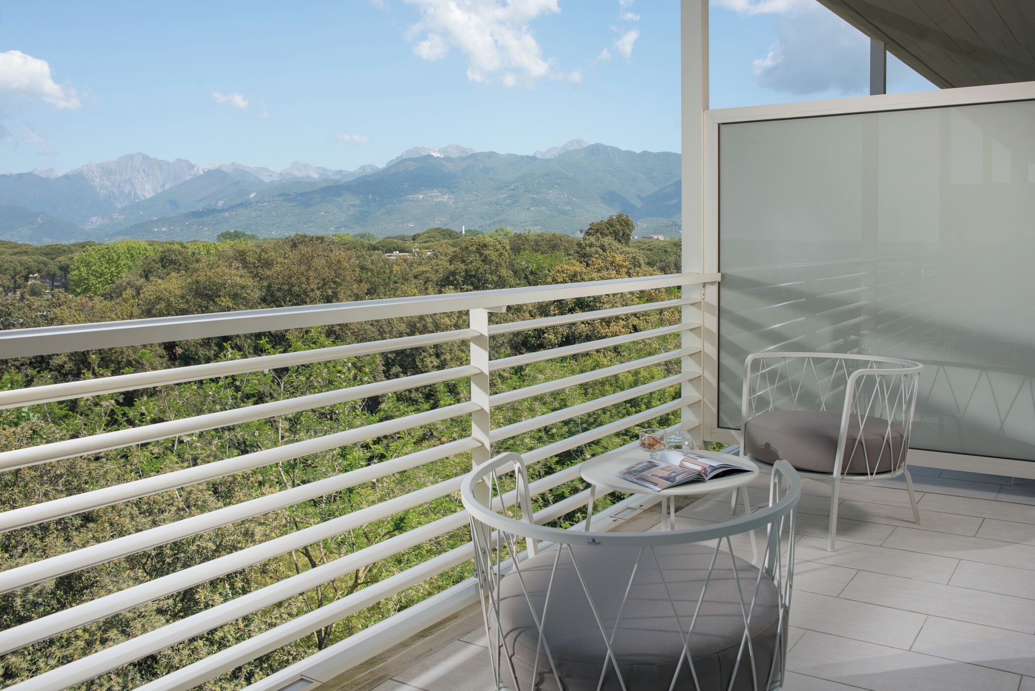 Een balkon met uitzicht op bergen, een ronde tafel met een stoel, en een glazen scheiding.