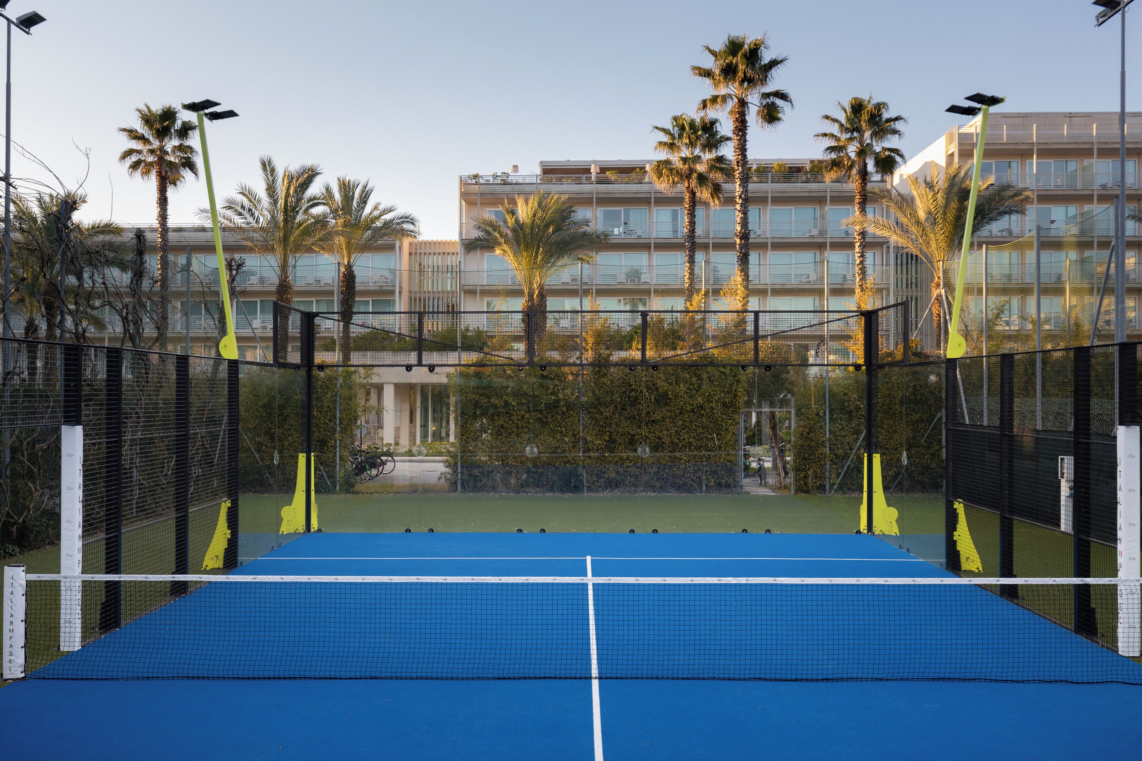 Ein Tennisplatz mit Netz, umgeben von einem Zaun. Im Hintergrund sind Palmen und ein Gebäude zu sehen.