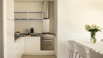 A modern kitchen with white cabinets, a stainless steel stove, and a coffee maker. There is a dining table with chairs and a vase of flowers.