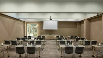 Une salle de conférence avec des rangées de tables et de chaises, un écran de projection et une grande fenêtre laissant entrer la lumière naturelle.