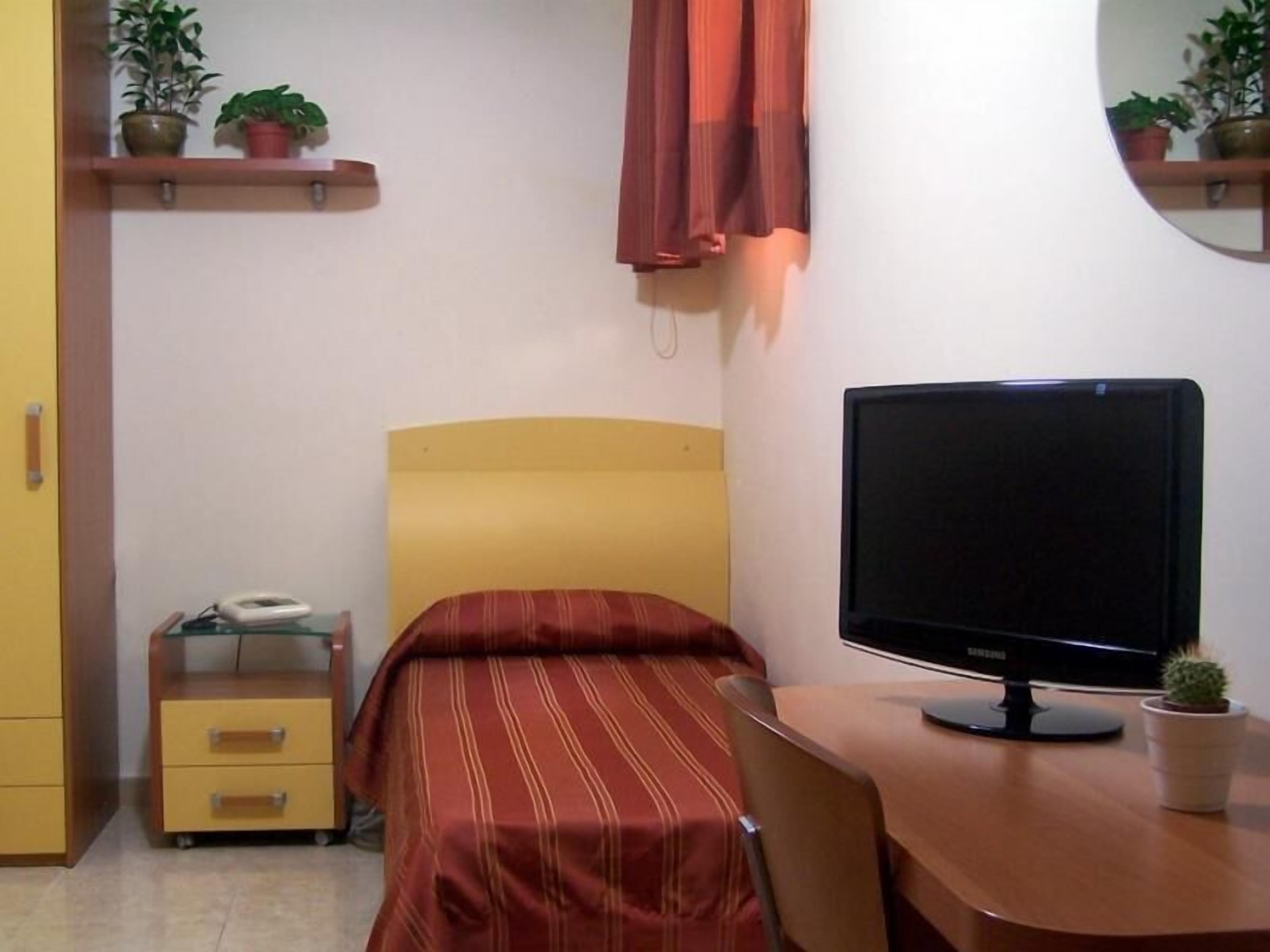 A bedroom with a bed, a desk with a computer, a chair, a bedside table with a phone, and a small potted plant.