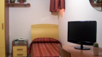 A bedroom with a bed, a desk with a computer, a chair, a bedside table with a phone, and a small potted plant.