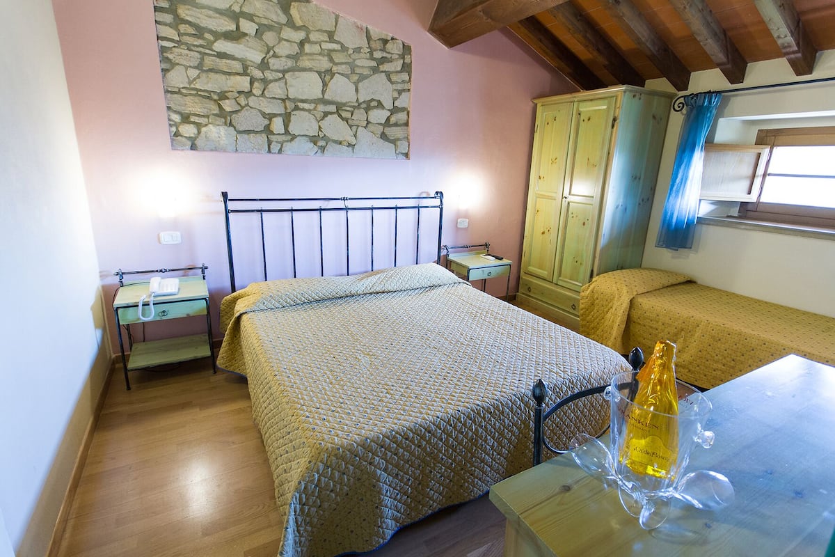 Un dormitorio con una cama, una mesita con una botella, una mesita de noche, un armario y una pared de piedra.